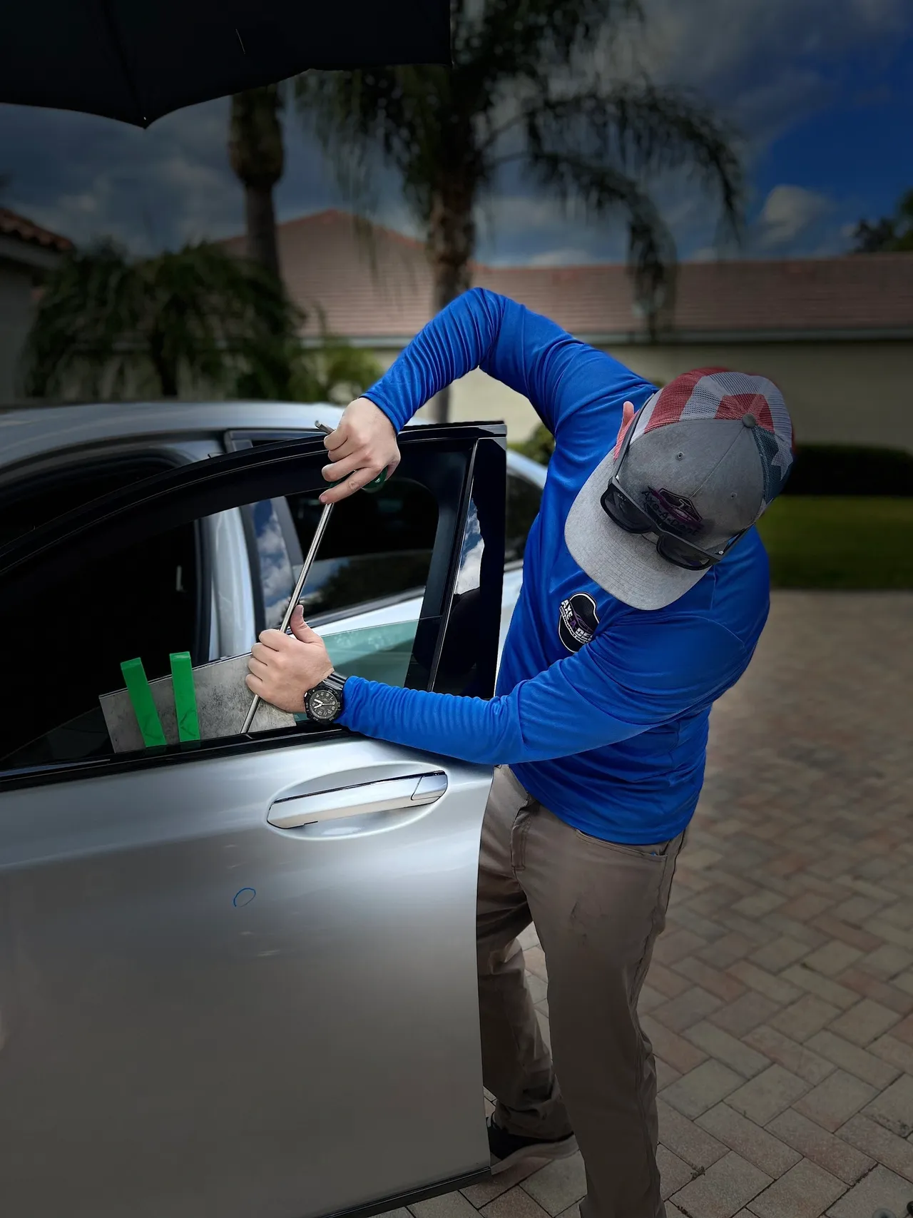 Man in blue shirt working on car window with tools outdoors.
