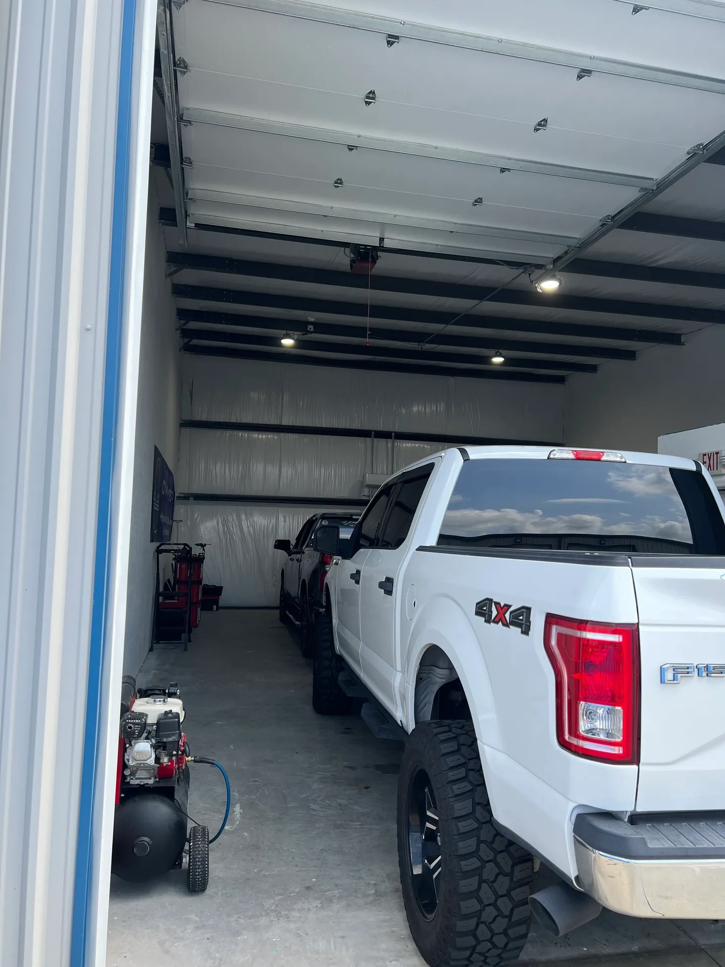 White pickup truck parked inside a garage, with another car visible. An air compressor sits near the entrance.