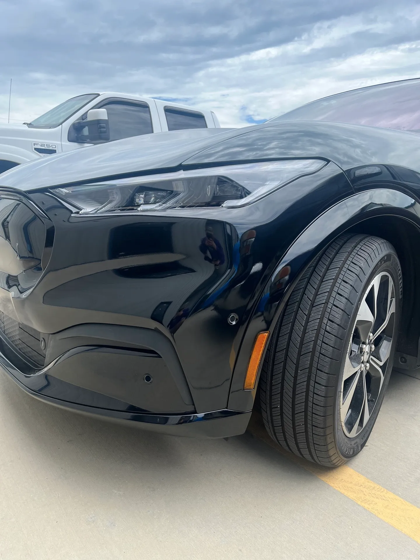 Black Ford Mustang Mach-E electric vehicle. Front corner view showing headlight, tire, and wheel.
