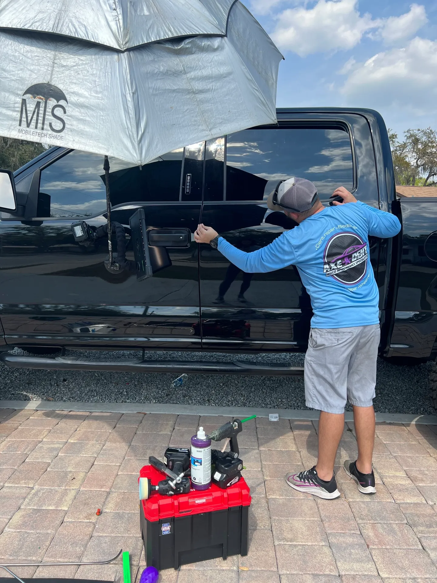 Person working on a black truck, using an umbrella. Tools are nearby. Outdoors, sunny.