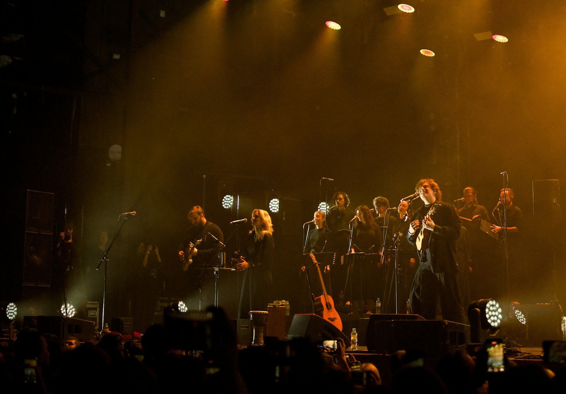 Band performing on a dark stage under warm golden spotlights, with audience silhouettes in front