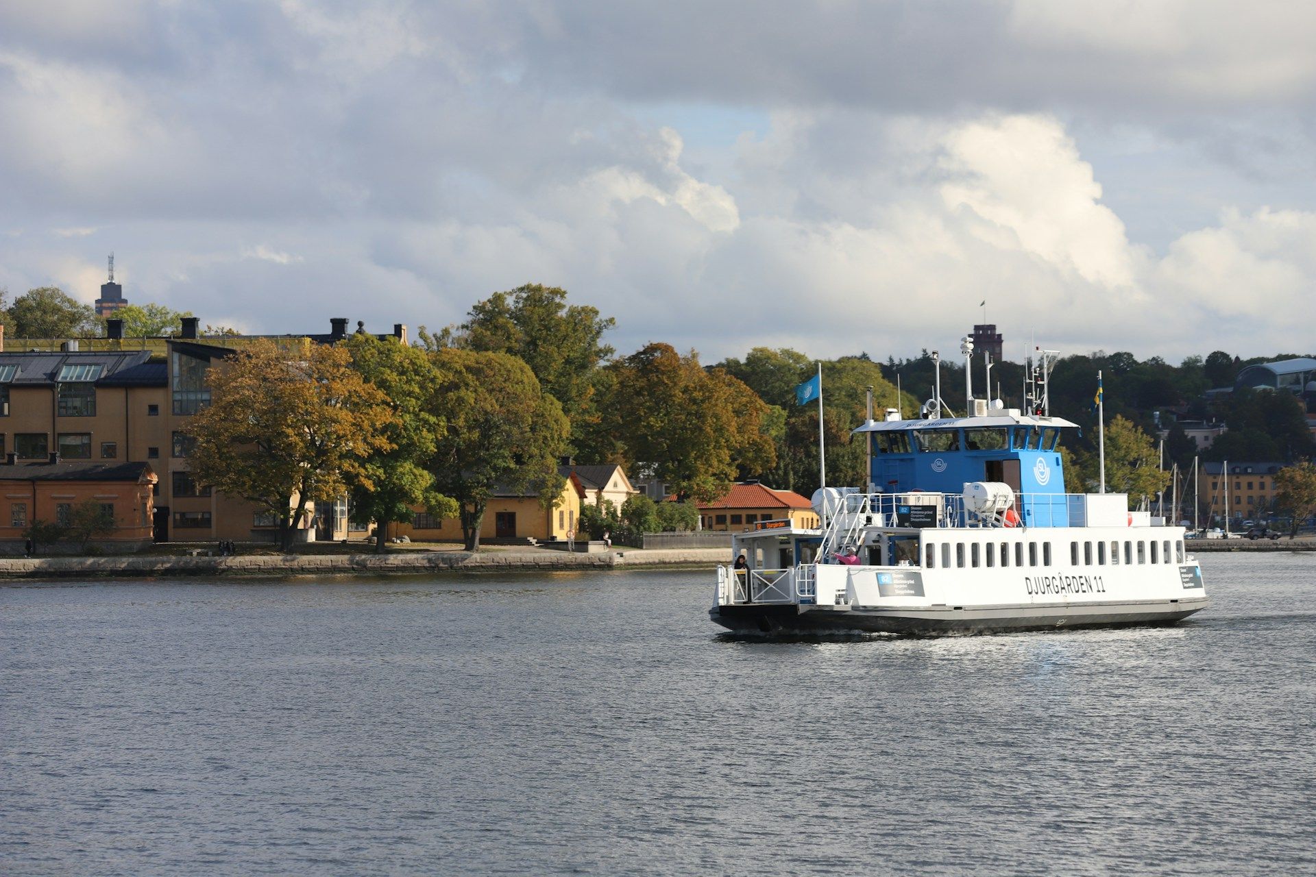 A passenger ferry travels across the water with Djurgården in the background.