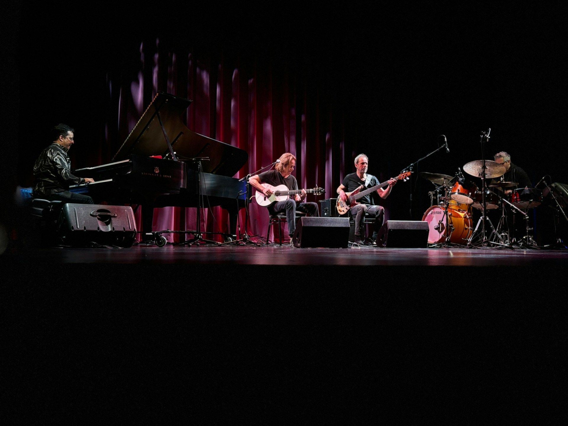 Jazz band performing on a dimly lit stage, with piano, guitar, bass, drums, and red curtains.