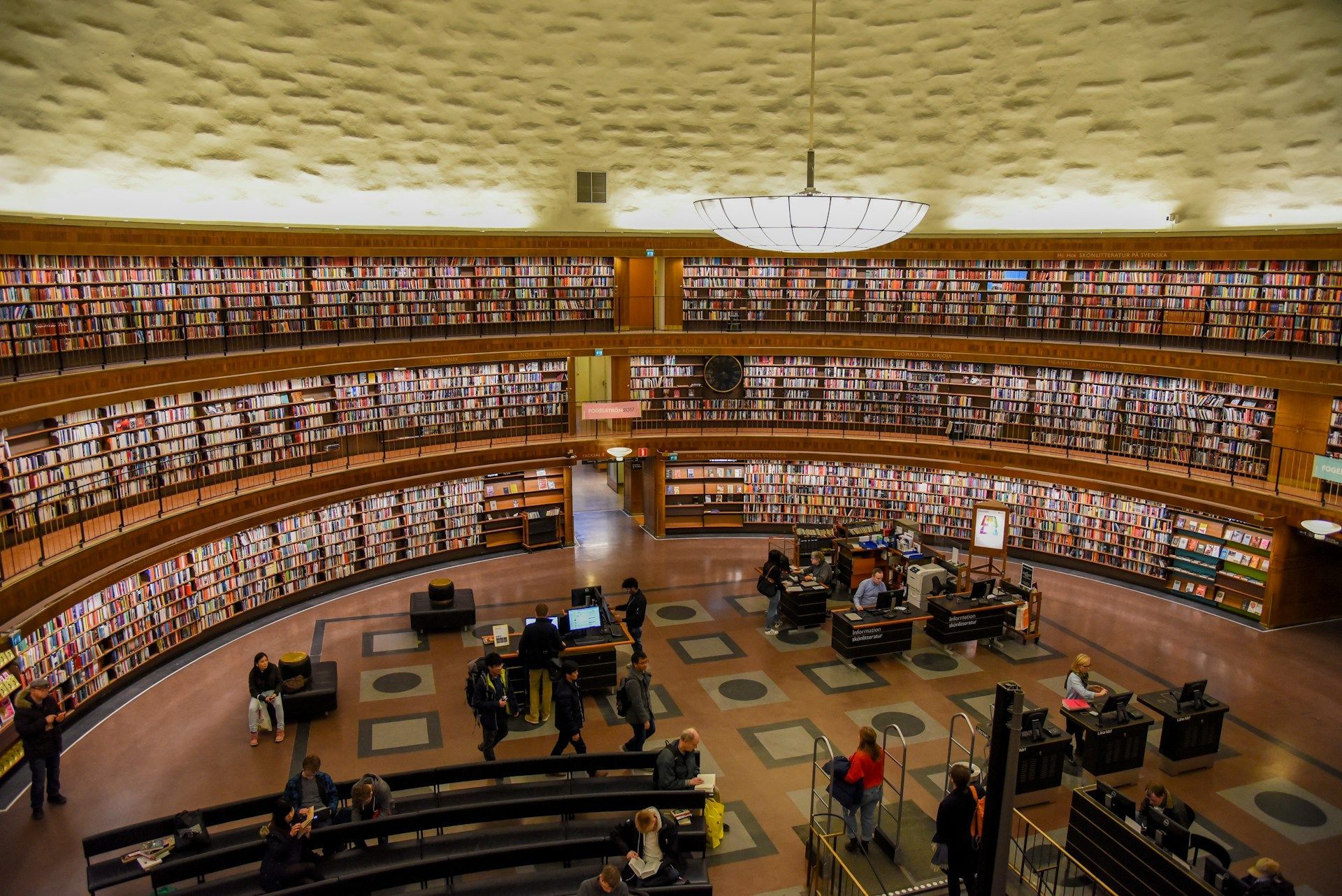 Grand library interior with tall bookshelves, curved balconies, and people at tables below