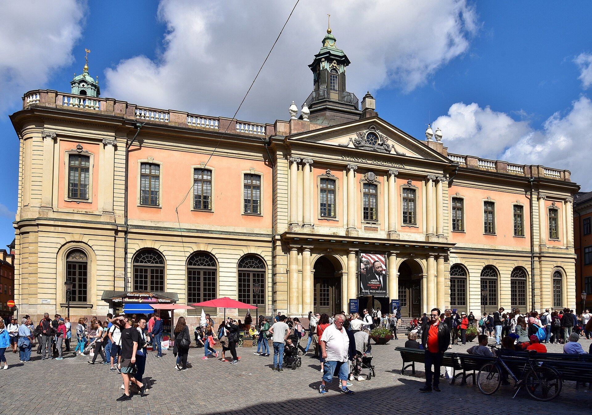 Crowded plaza before a historic pale pink and beige building under a blue sky