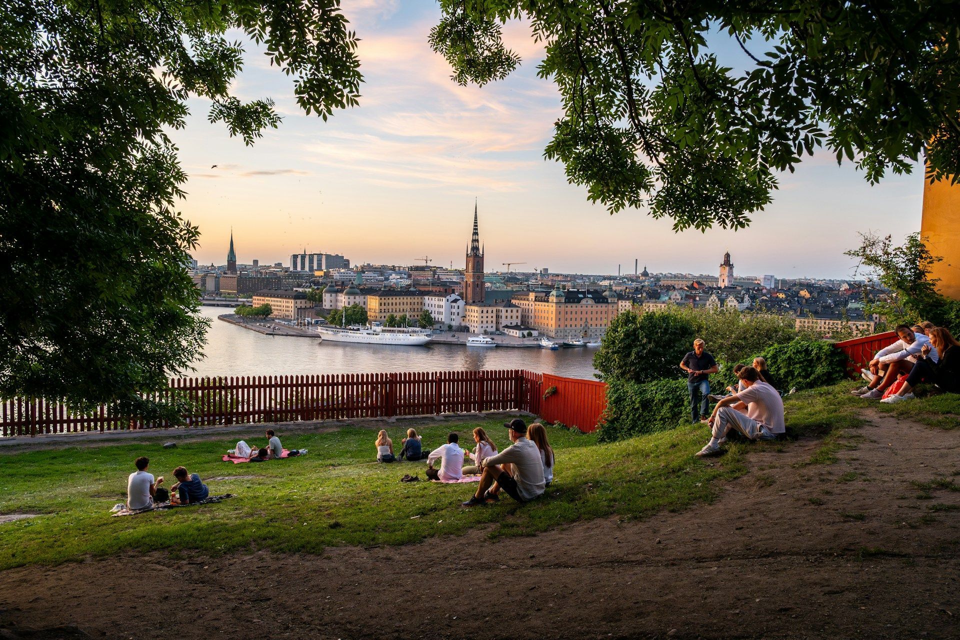 People sit on a grassy hill at Södermalm overlooking the Stockholm waterfront cityscape during sunset.