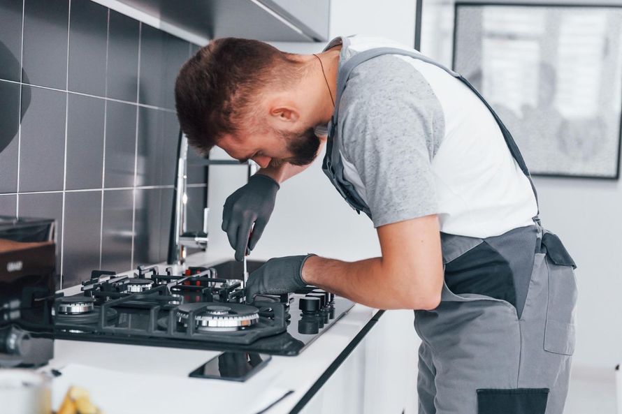 Technician repairing a gas stove in a modern kitchen, wearing gloves and leaning over the cooktop.
