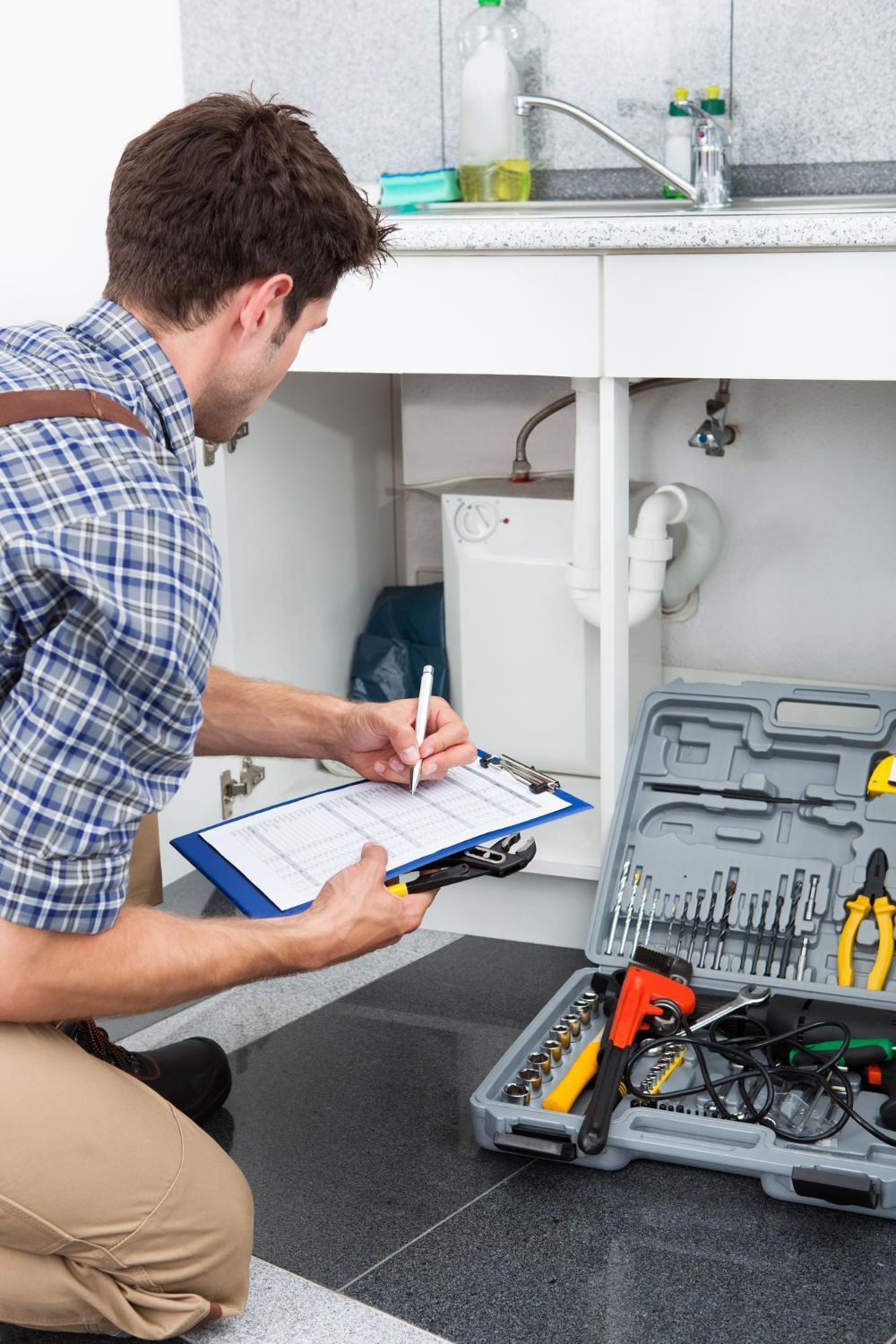 Plumber inspecting under-sink pipes in a kitchen with tools open on the floor.