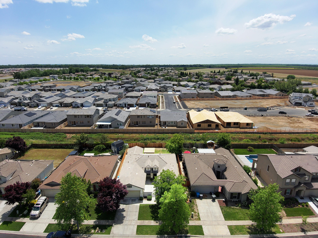 Aerial view of a suburban neighborhood with rows of houses, streets, and open fields under a blue sky