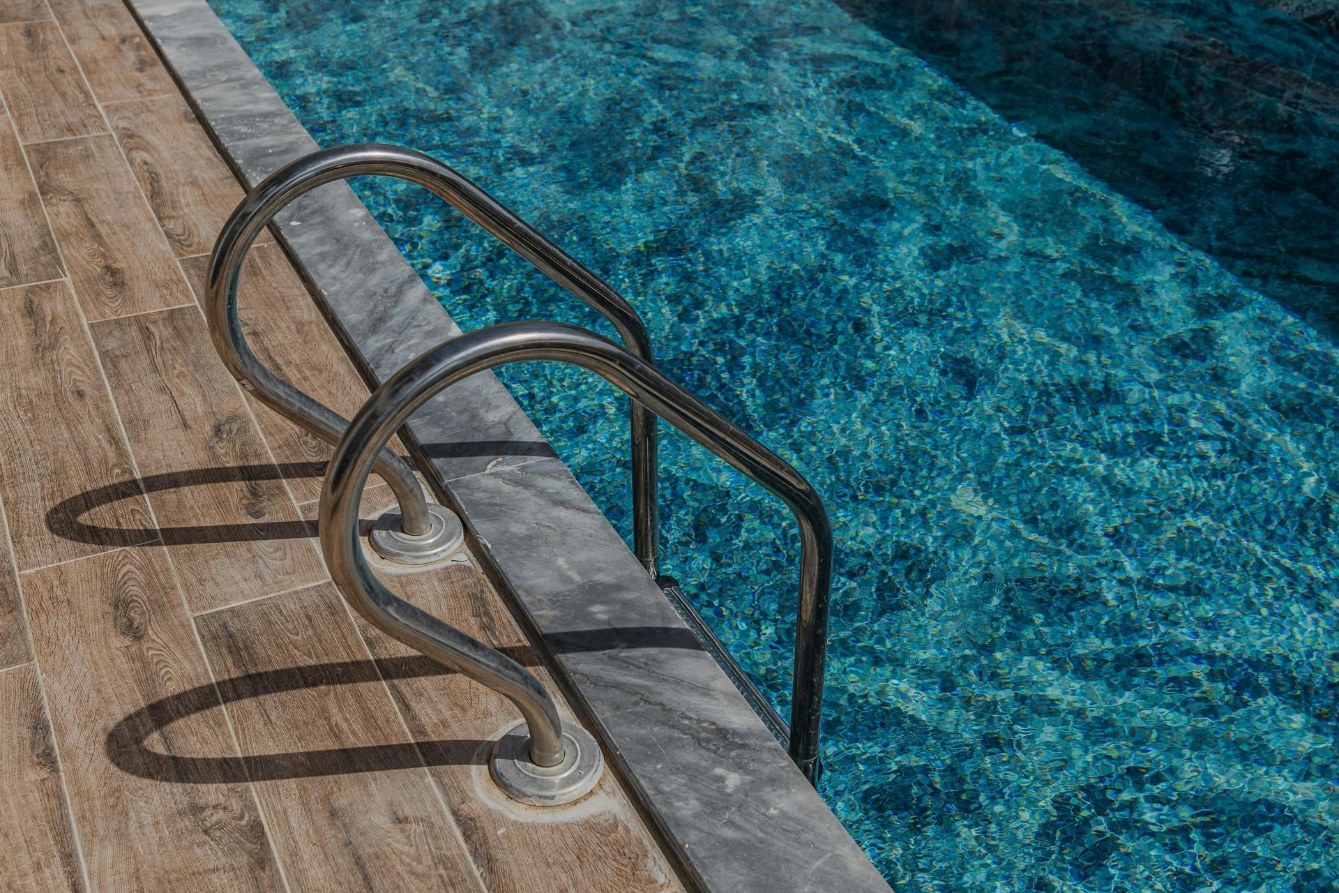 Pool ladder beside blue water and weathered wooden deck
