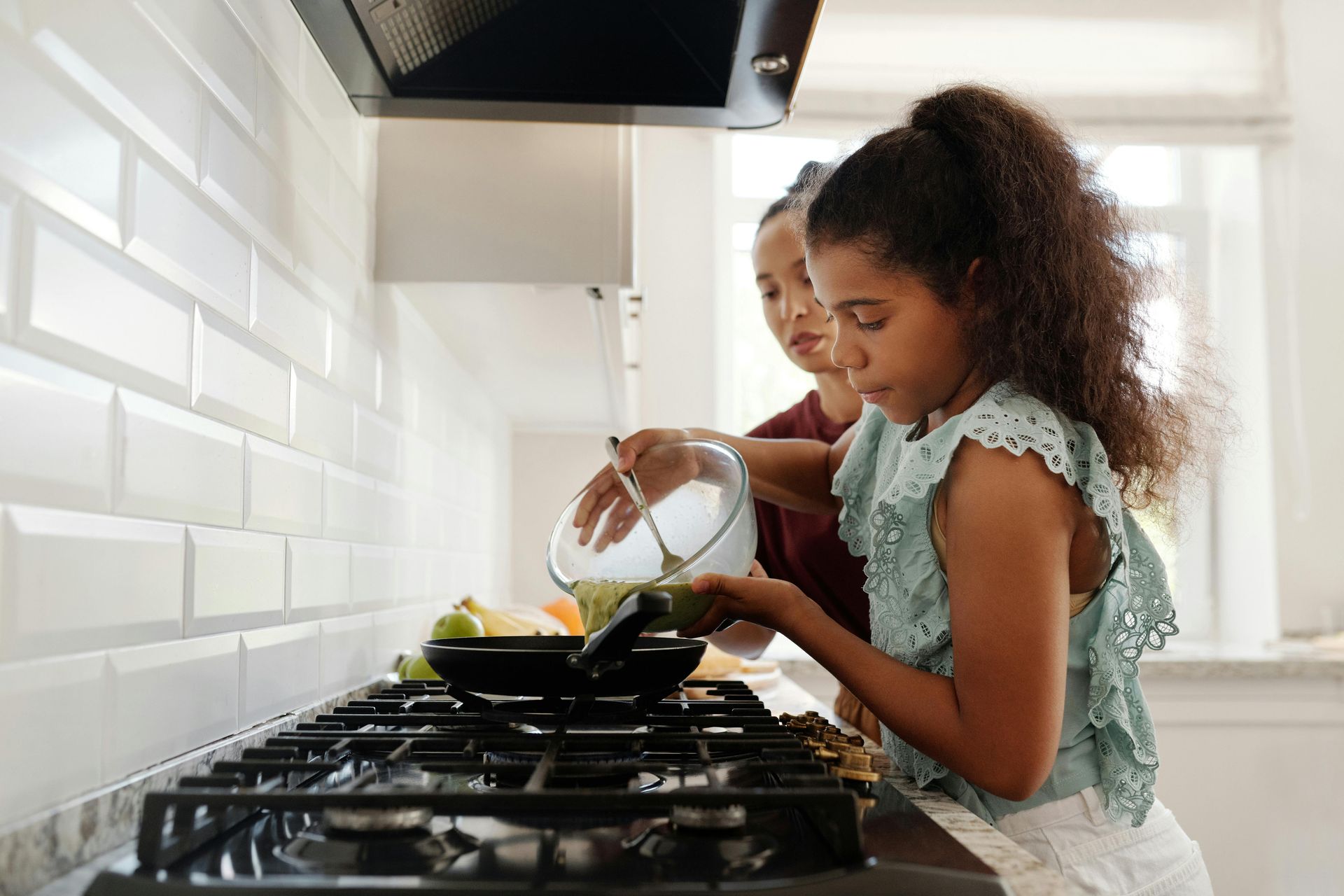 Child pouring ingredients into a pan on a stove in a bright kitchen