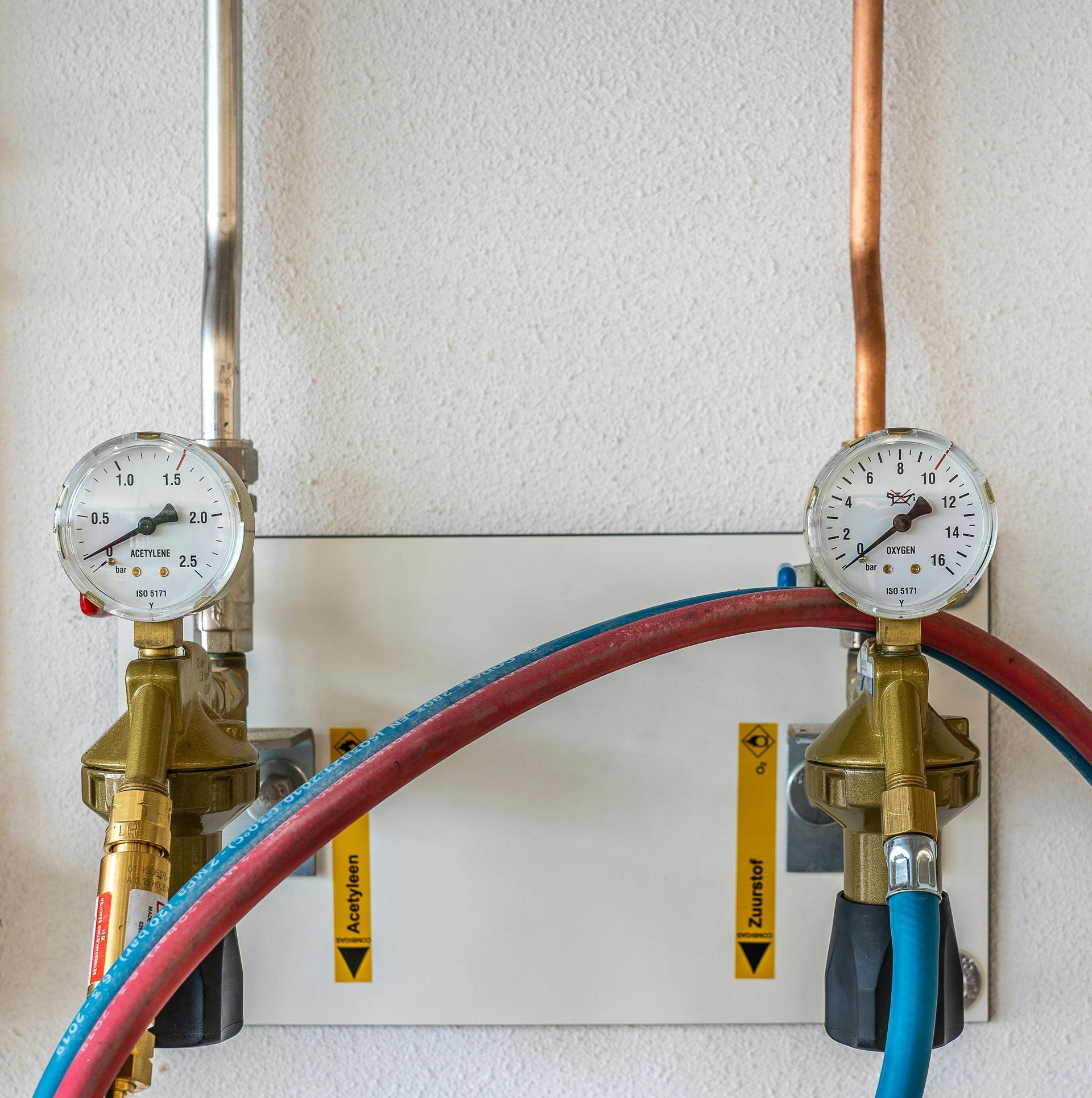 Two wall-mounted gauges on brass valves with red and blue hoses in a mechanical room
