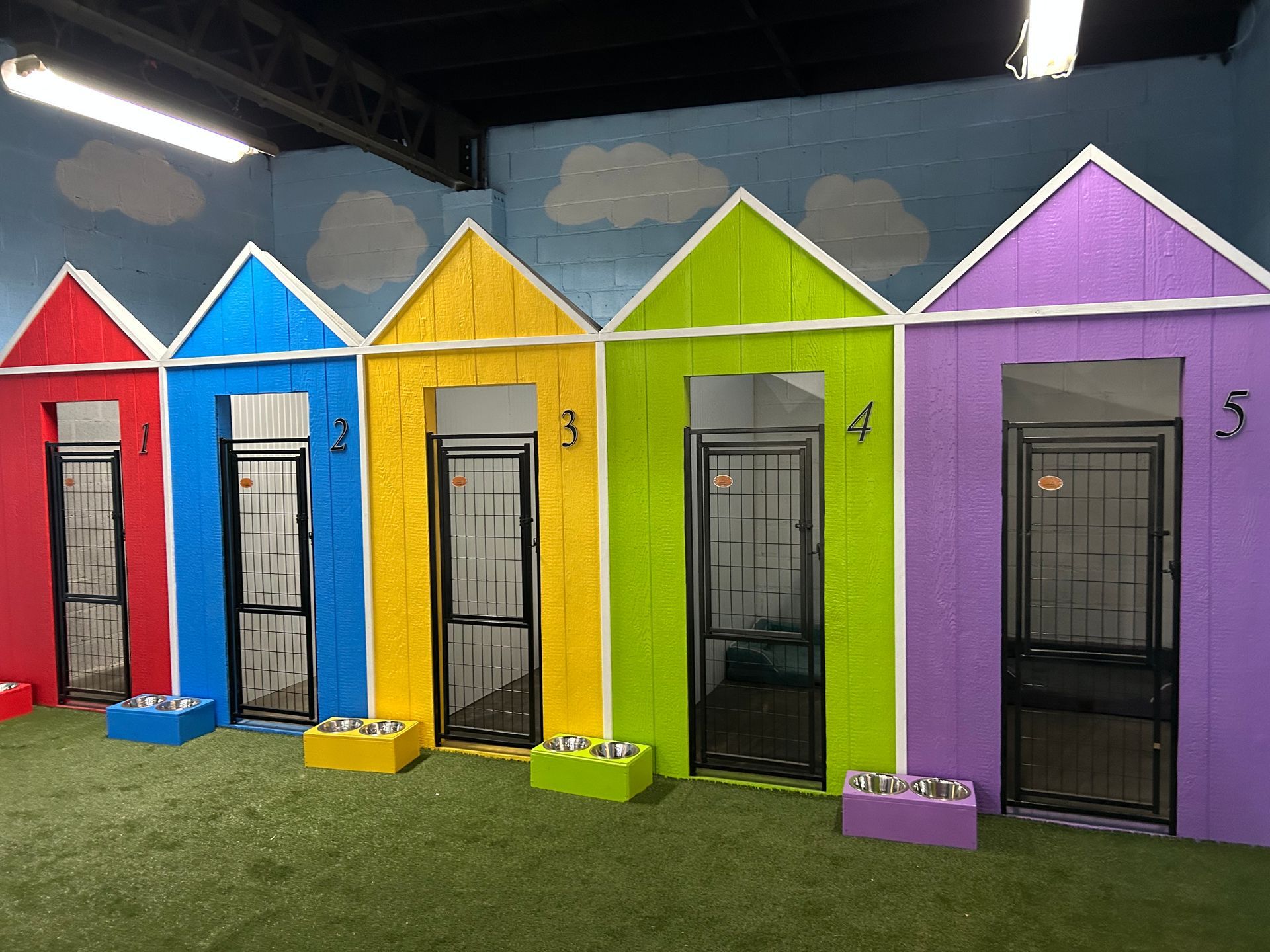 Row of colorful dog kennels, each with a different color, numbered 1-5, in an indoor play area.