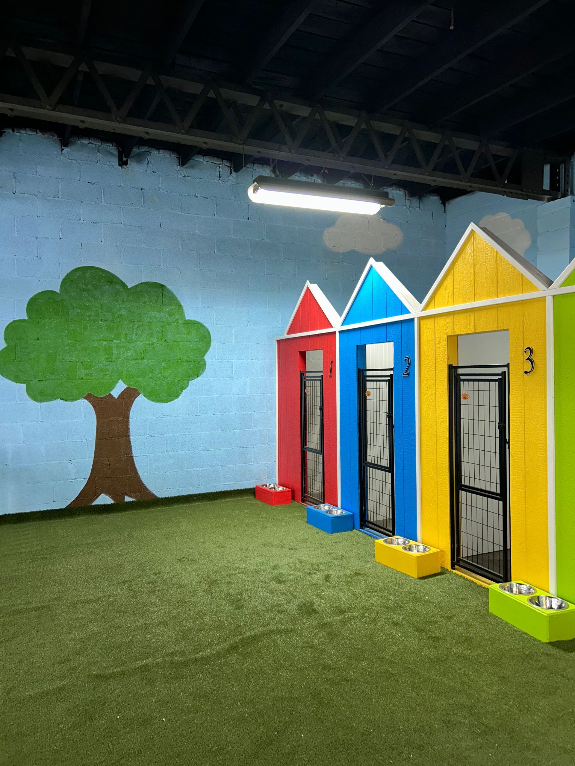 Colorful dog kennels with house-shaped roofs, a painted tree, and artificial turf.