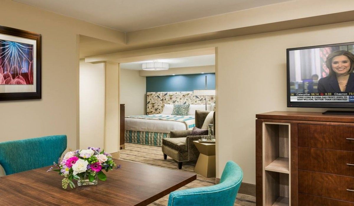 Hotel suite with teal chairs, wooden table, TV, and a bedroom visible through an open doorway