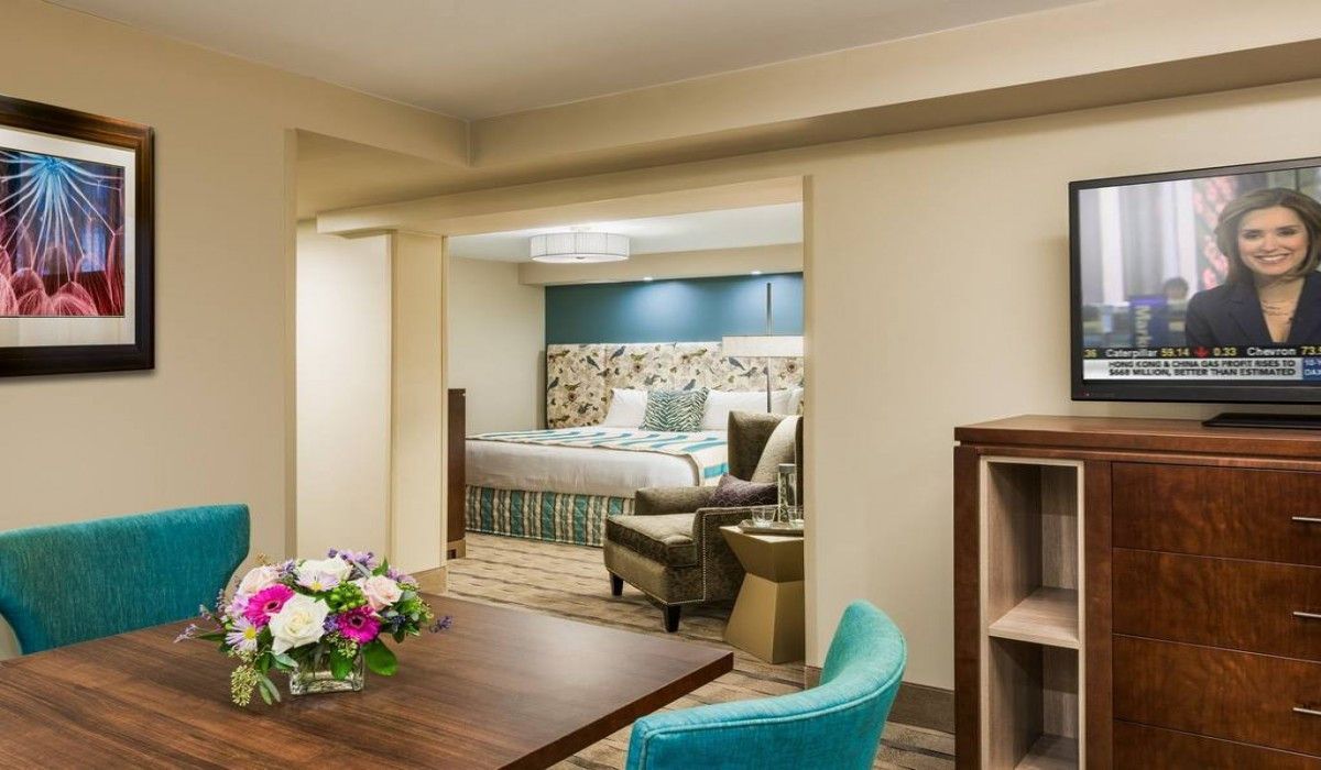 Hotel room suite with dining table, teal chairs, bedroom alcove, and TV on a wooden stand