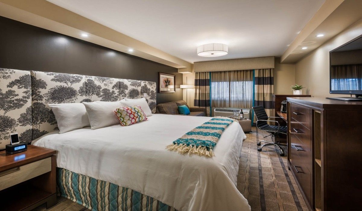 Modern hotel room with a king bed, patterned headboard, desk, TV, and city view through curtains