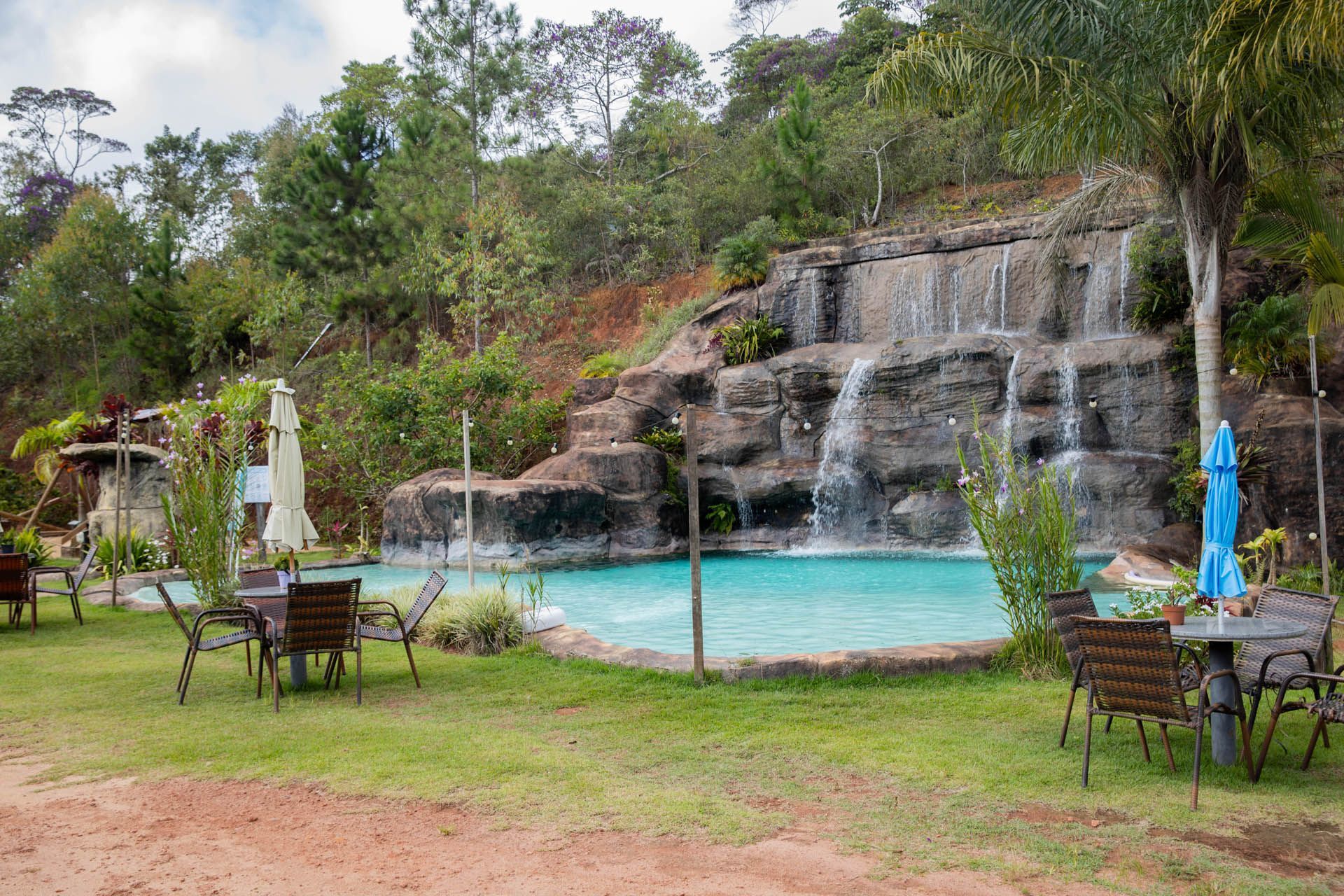 Piscina no jardim com cascata rochosa, água turquesa e mesas de pátio com guarda-sóis.