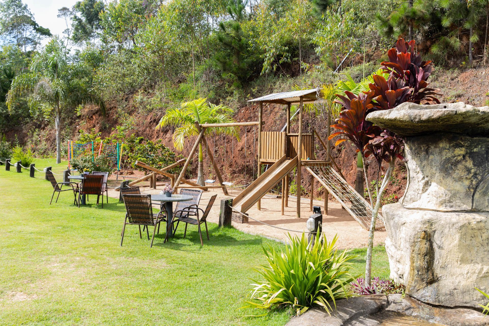 Jardim nos fundos com mesas de pátio, balanço e fonte de pedra em um ambiente verdejante.