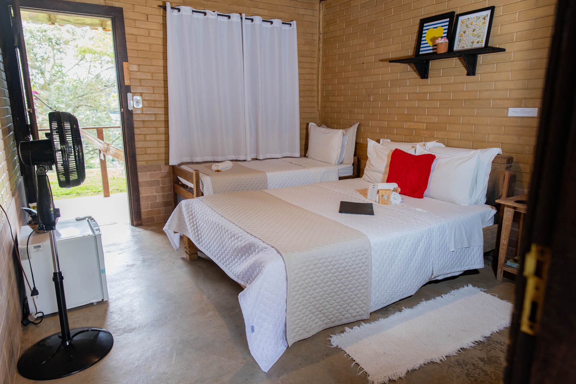 Quarto aconchegante com duas camas brancas, paredes de tijolos aparentes, cortina na janela e ventilador de pé junto à porta.