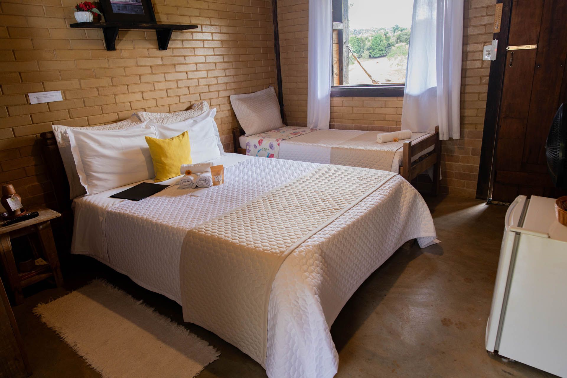 Quarto aconchegante com duas camas, paredes de tijolos aparentes, cortinas brancas e uma janela que deixa entrar a luz do dia.
