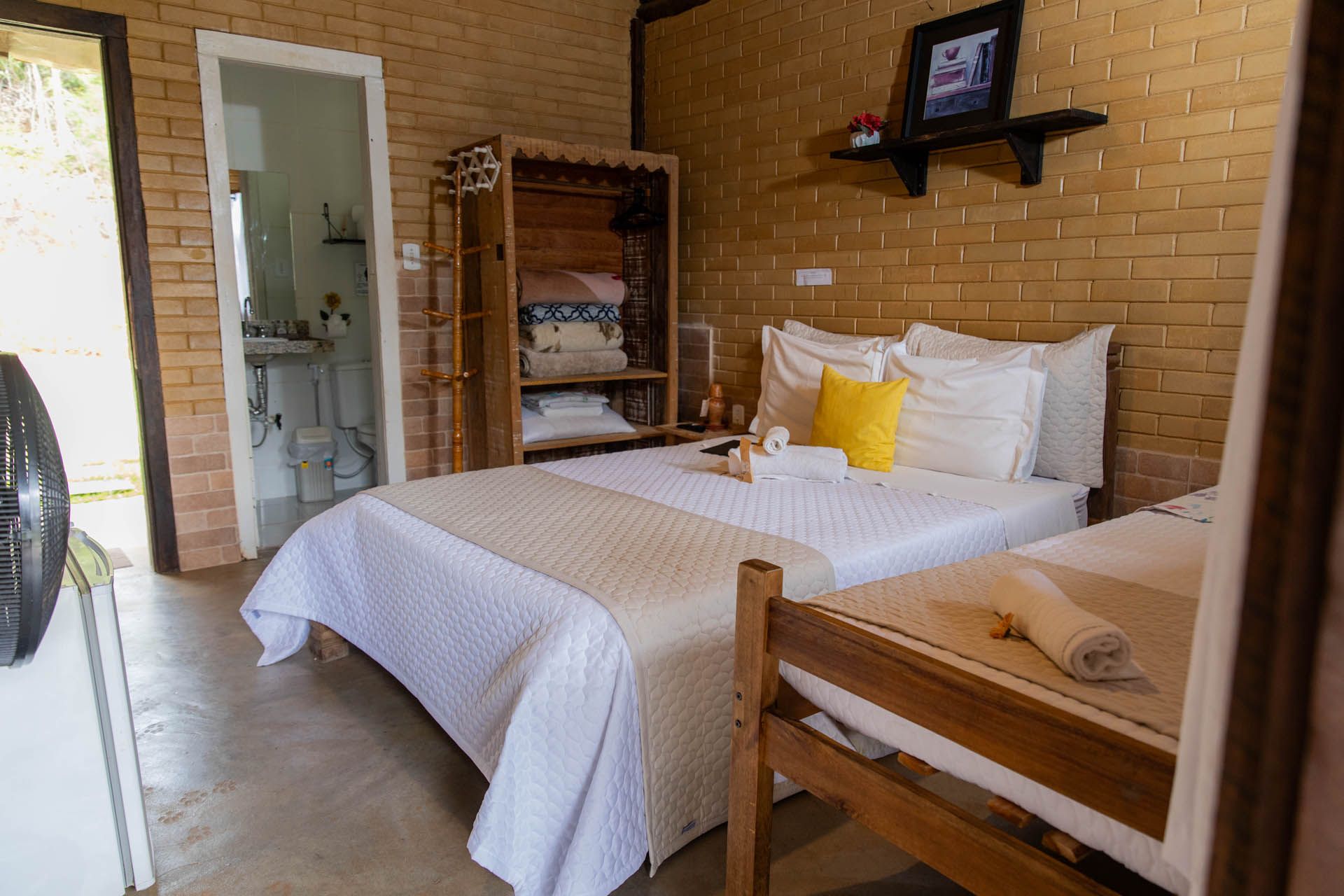 Quarto aconchegante com colcha branca, móveis de madeira, parede de tijolos aparentes e porta do banheiro aberta.
