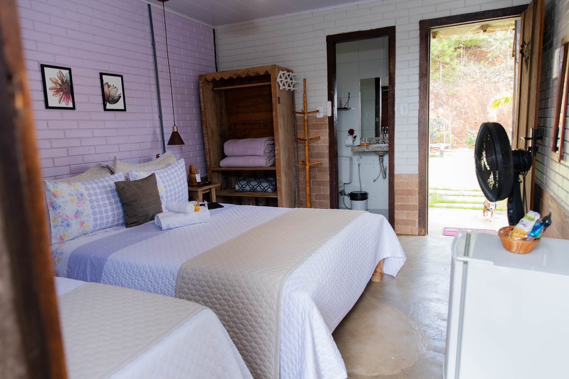 Quarto aconchegante com duas camas de solteiro, paredes cor lavanda e uma porta aberta para um pátio iluminado.