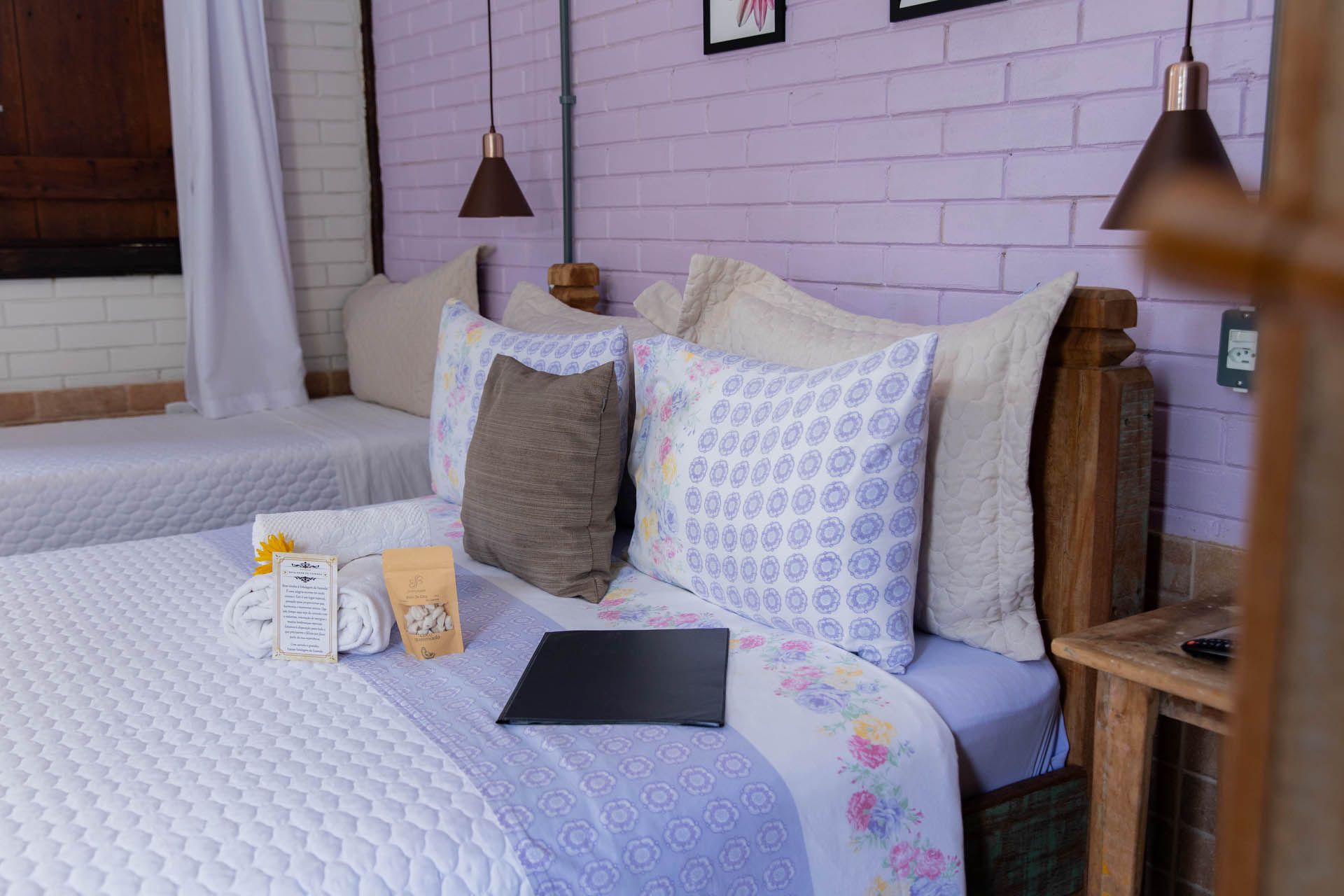 Cama arrumada com colcha branca, almofadas estampadas e um laptop em um quarto aconchegante com uma parede de tijolos rosa.