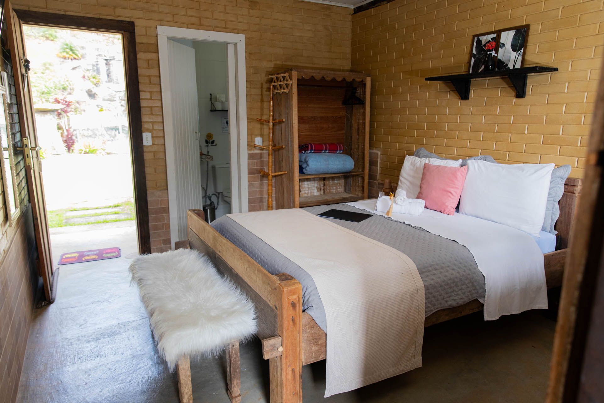 Quarto aconchegante com cama de madeira, roupa de cama cinza, travesseiro rosa, banco de pele sintética e porta aberta para uma varanda iluminada.