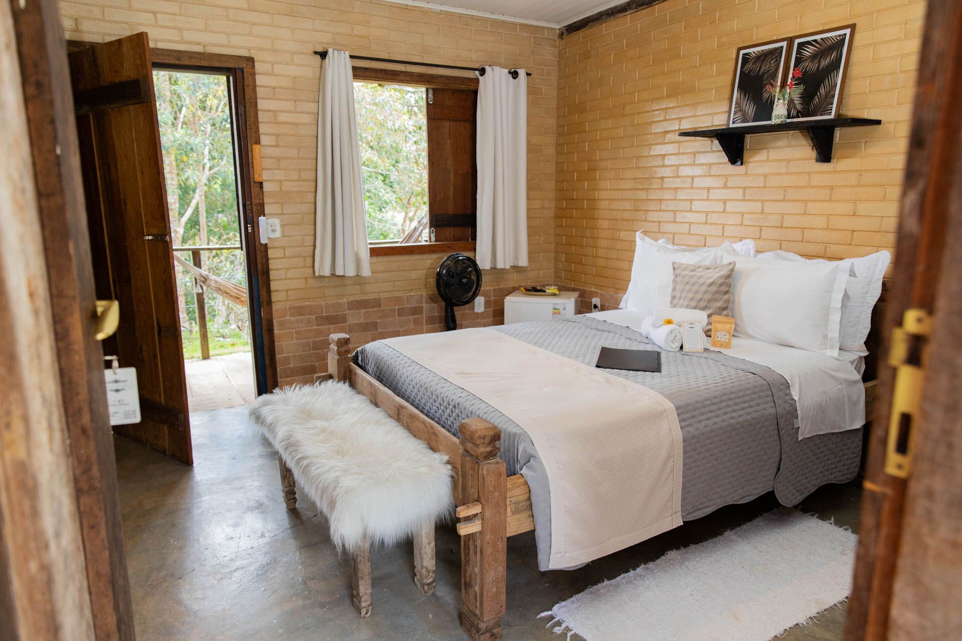 Quarto rústico e aconchegante com uma cama grande, lençóis brancos, banco, porta aberta e luz natural entrando pelas janelas.