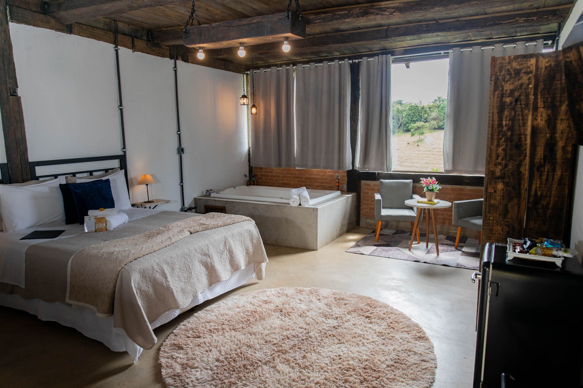 Quarto aconchegante com cama, tapete redondo, sofá, escrivaninha e janela grande com cortinas.