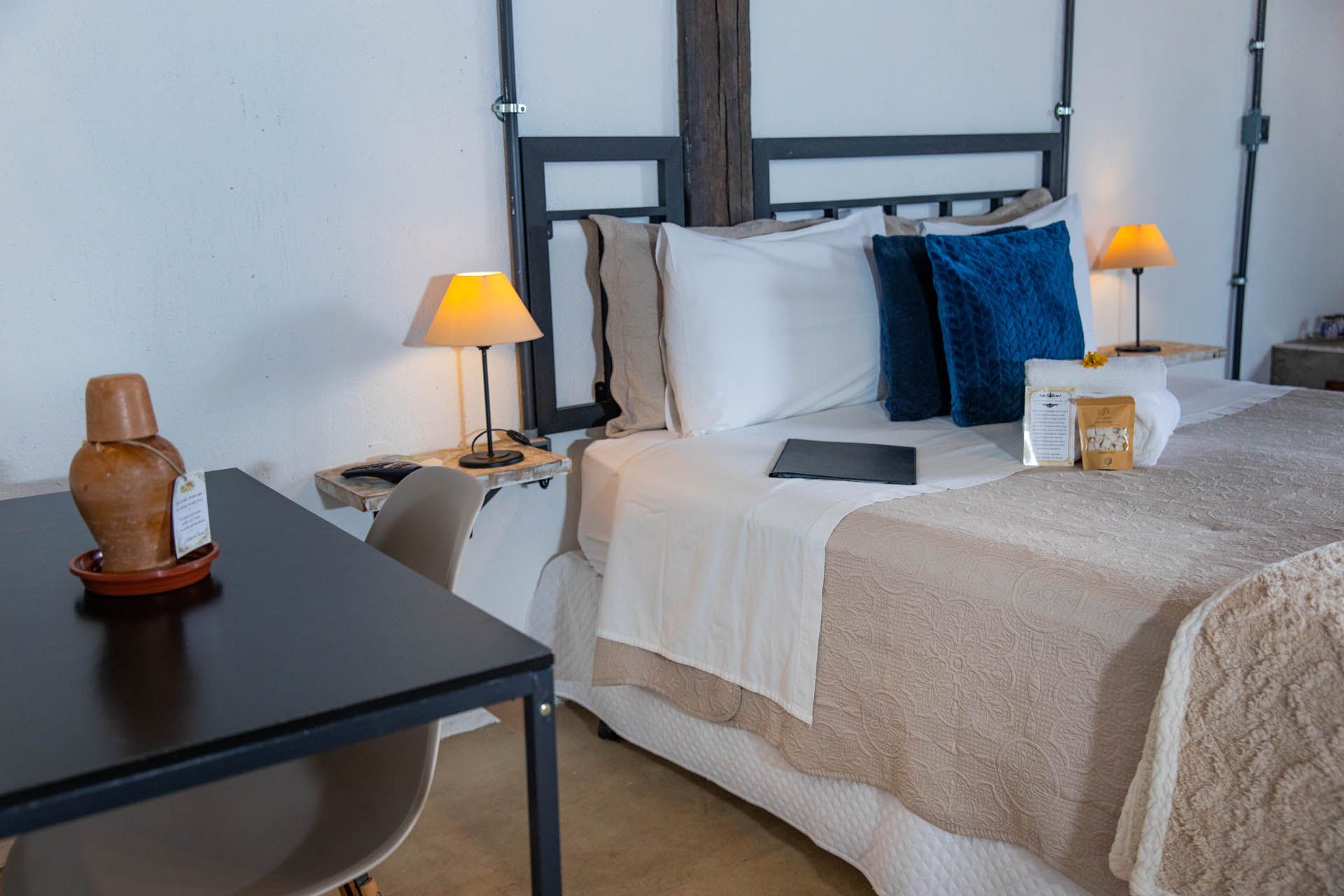 Cama arrumada num quarto luminoso com candeeiros de cabeceira e uma secretária preta em primeiro plano.
