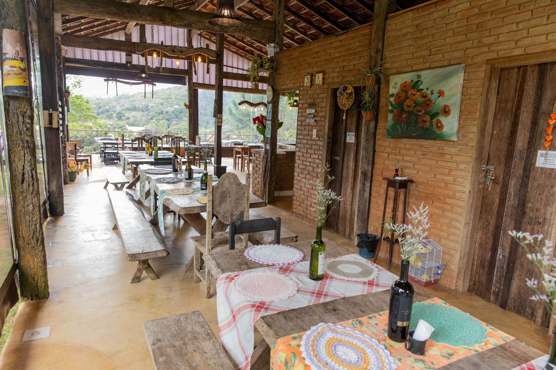 Restaurante rústico ao ar livre com mesas de madeira, paredes de tijolos e decoração floral com vista para um jardim.