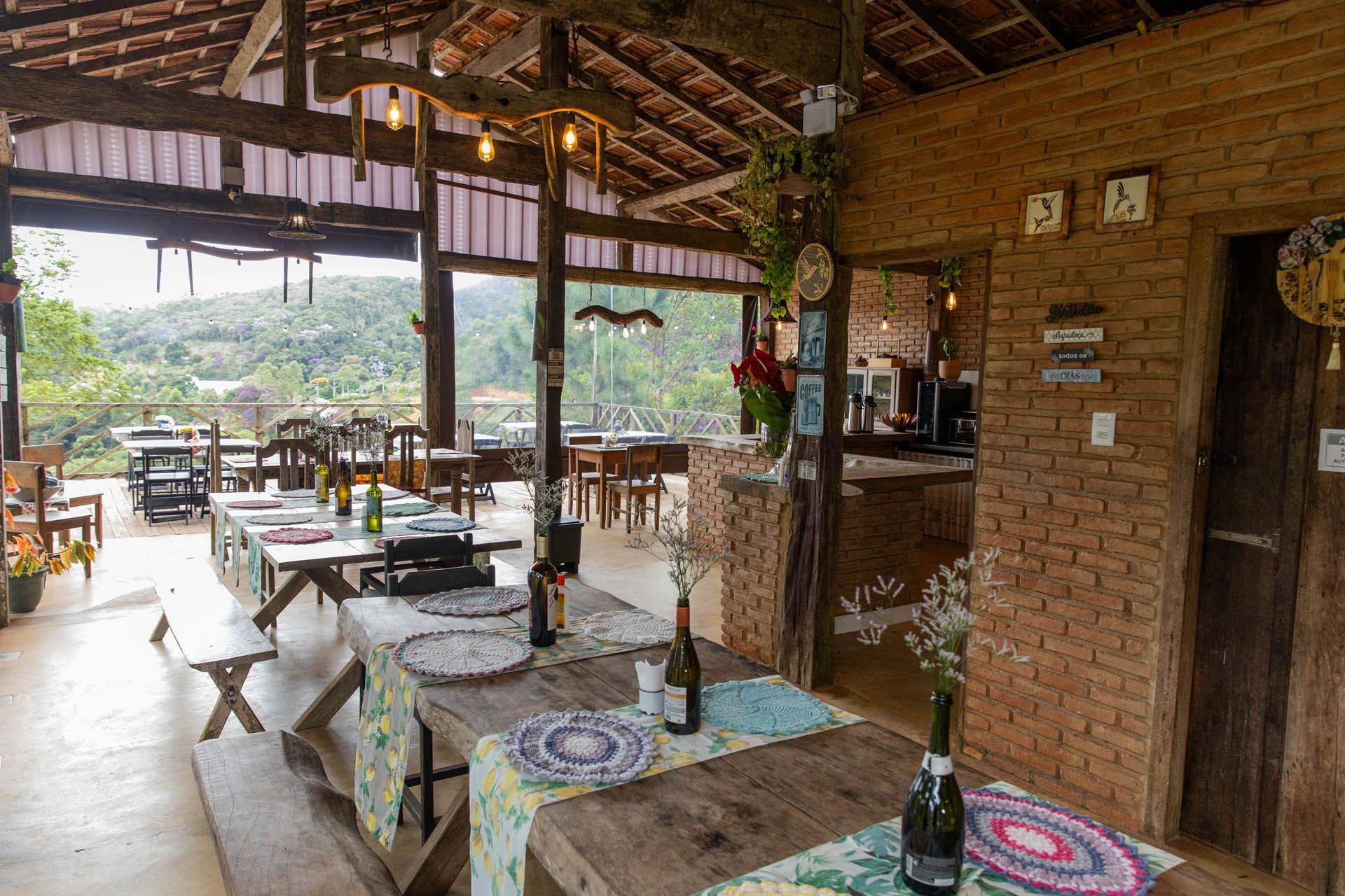 Restaurante rústico ao ar livre com mesas sob um telhado de palha, com vista para uma encosta verdejante.