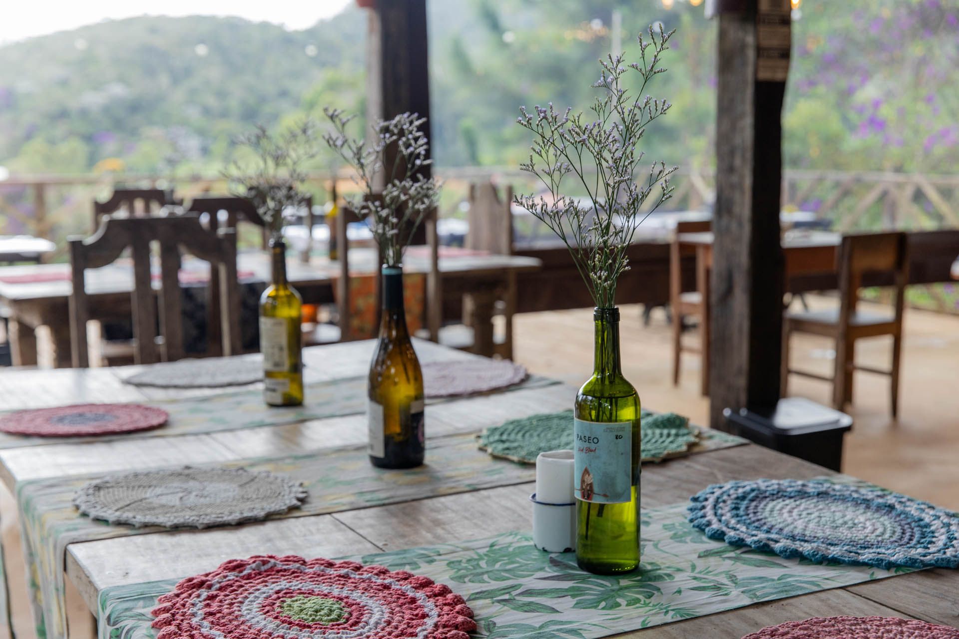 Mesas de café ao ar livre com jogos americanos de palha e garrafas verdes com vista para uma montanha desfocada.