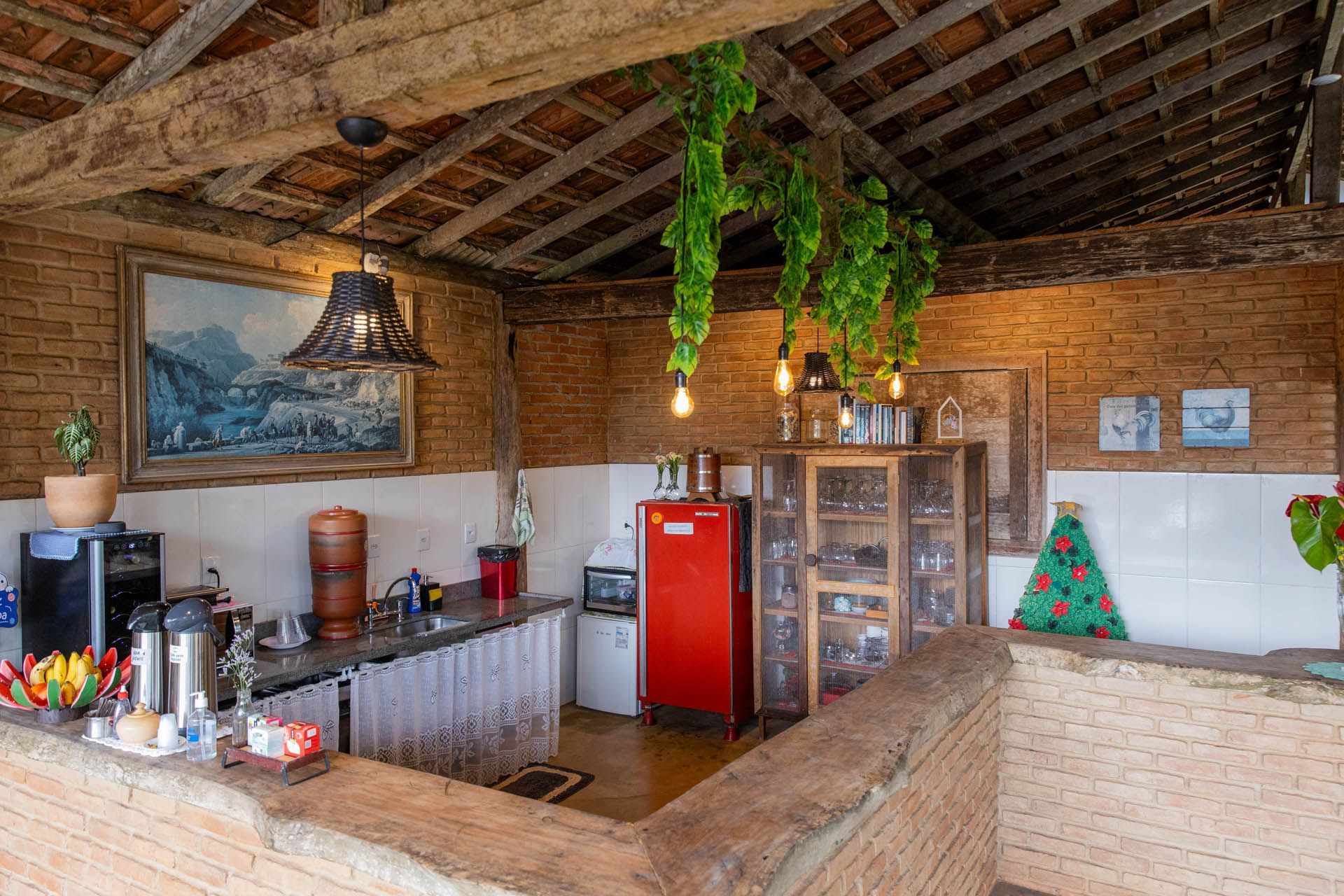Cozinha rústica com paredes de tijolos, vigas de madeira, geladeira vermelha, plantas suspensas e balcão de bar.
