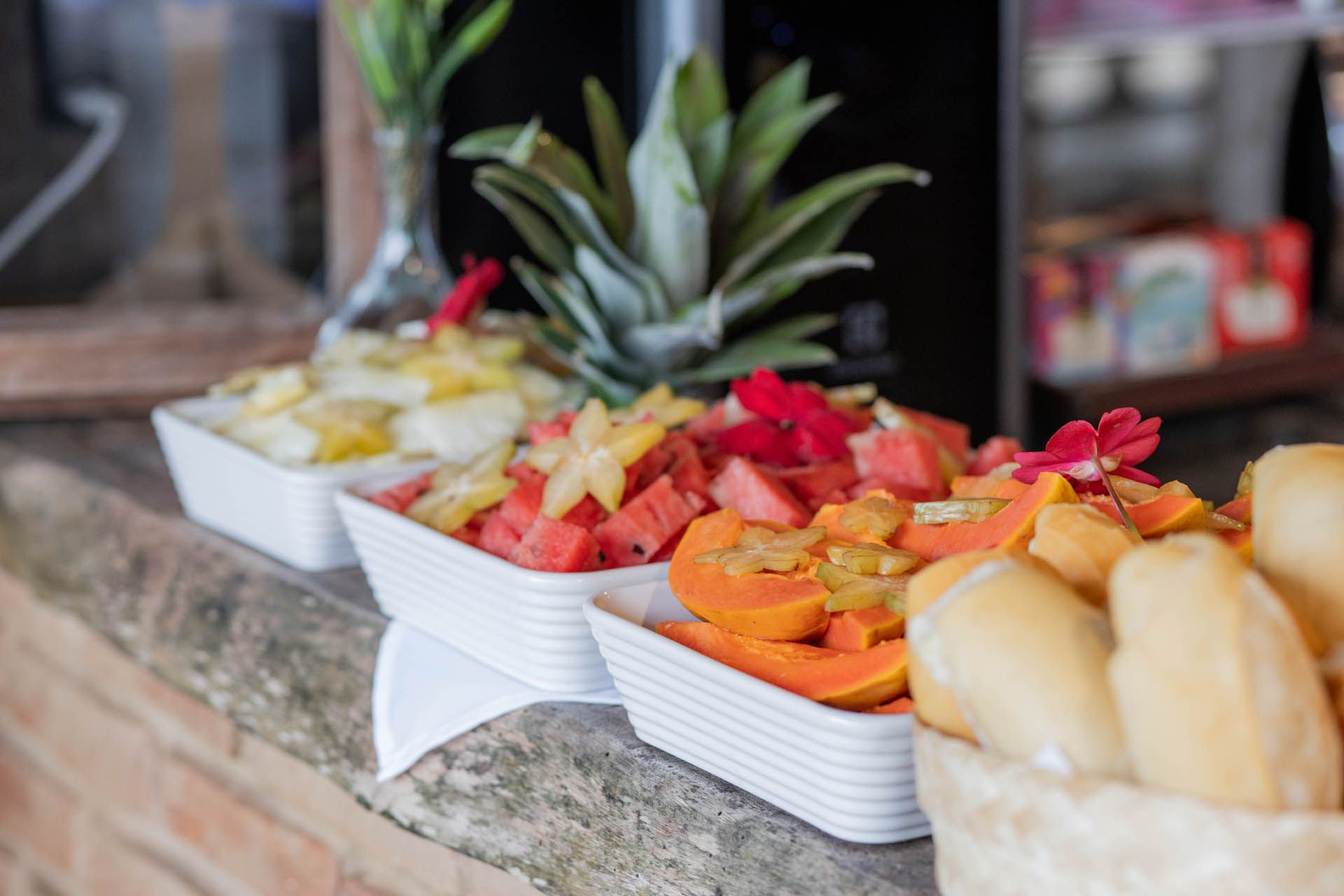 Pratos com diversas frutas sobre uma bancada de granito, incluindo abacaxi, melancia, frutas cítricas e pãezinhos.