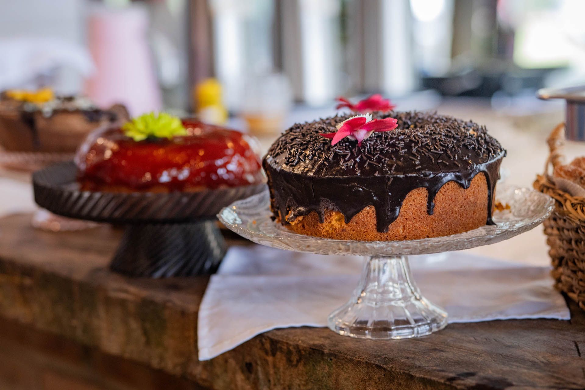 Bolo com cobertura de chocolate em um suporte de vidro ao lado de um bolo vermelho brilhante em uma mesa de madeira.