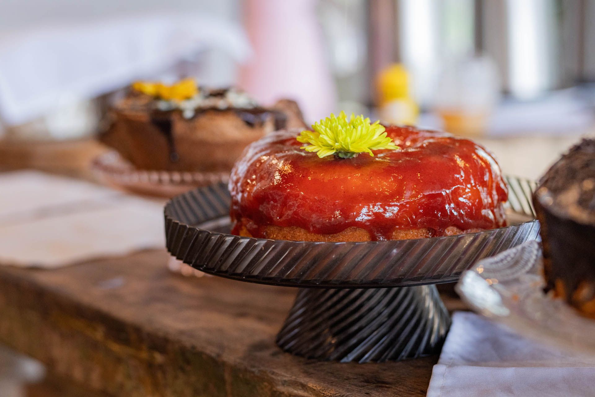 Tarte com cobertura de chocolate e flor amarela sobre uma mesa de madeira, com outros doces desfocados ao fundo.