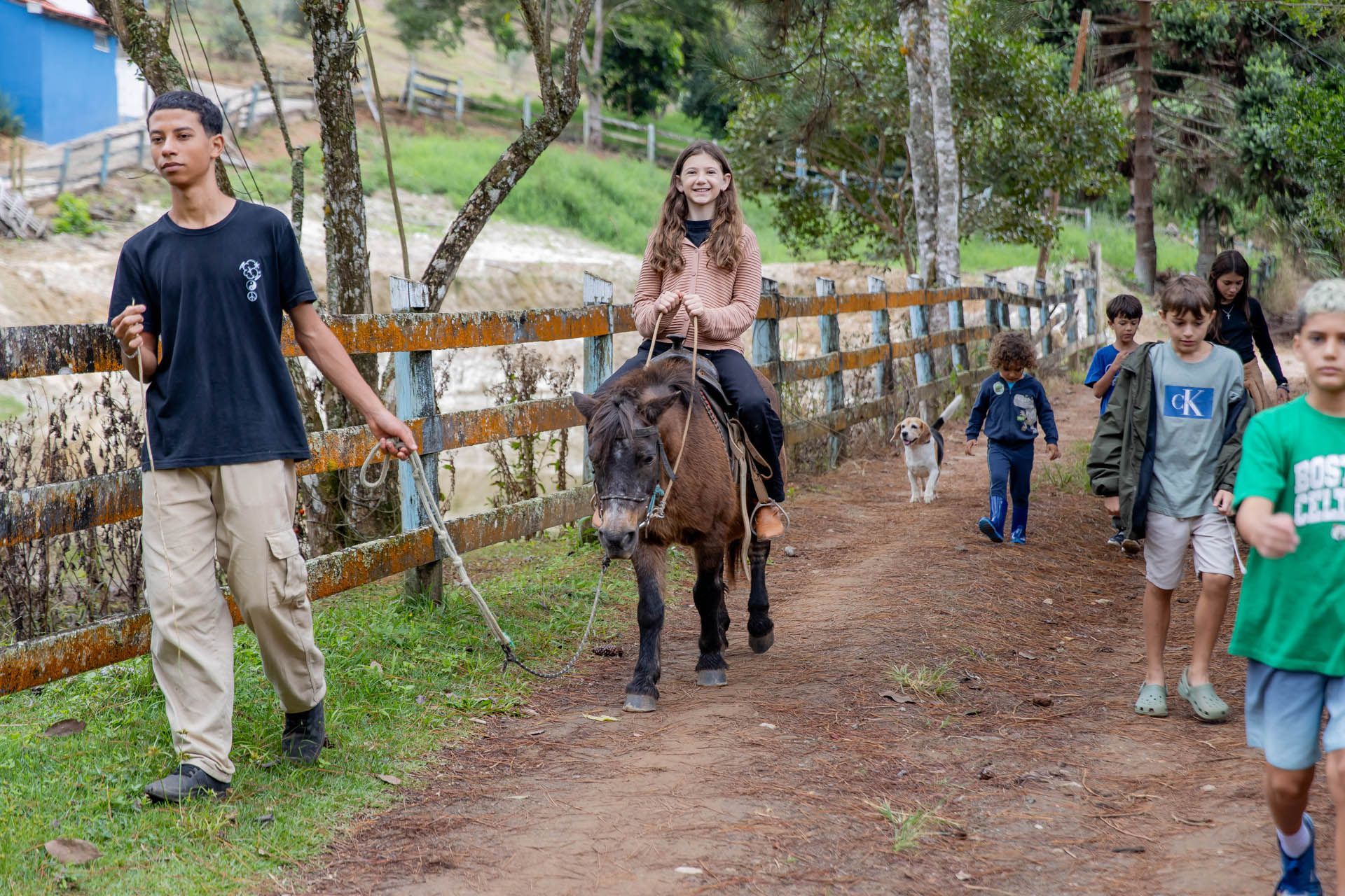 Pessoa montando um burro em uma trilha de terra, com várias pessoas caminhando ao lado em um parque rural.