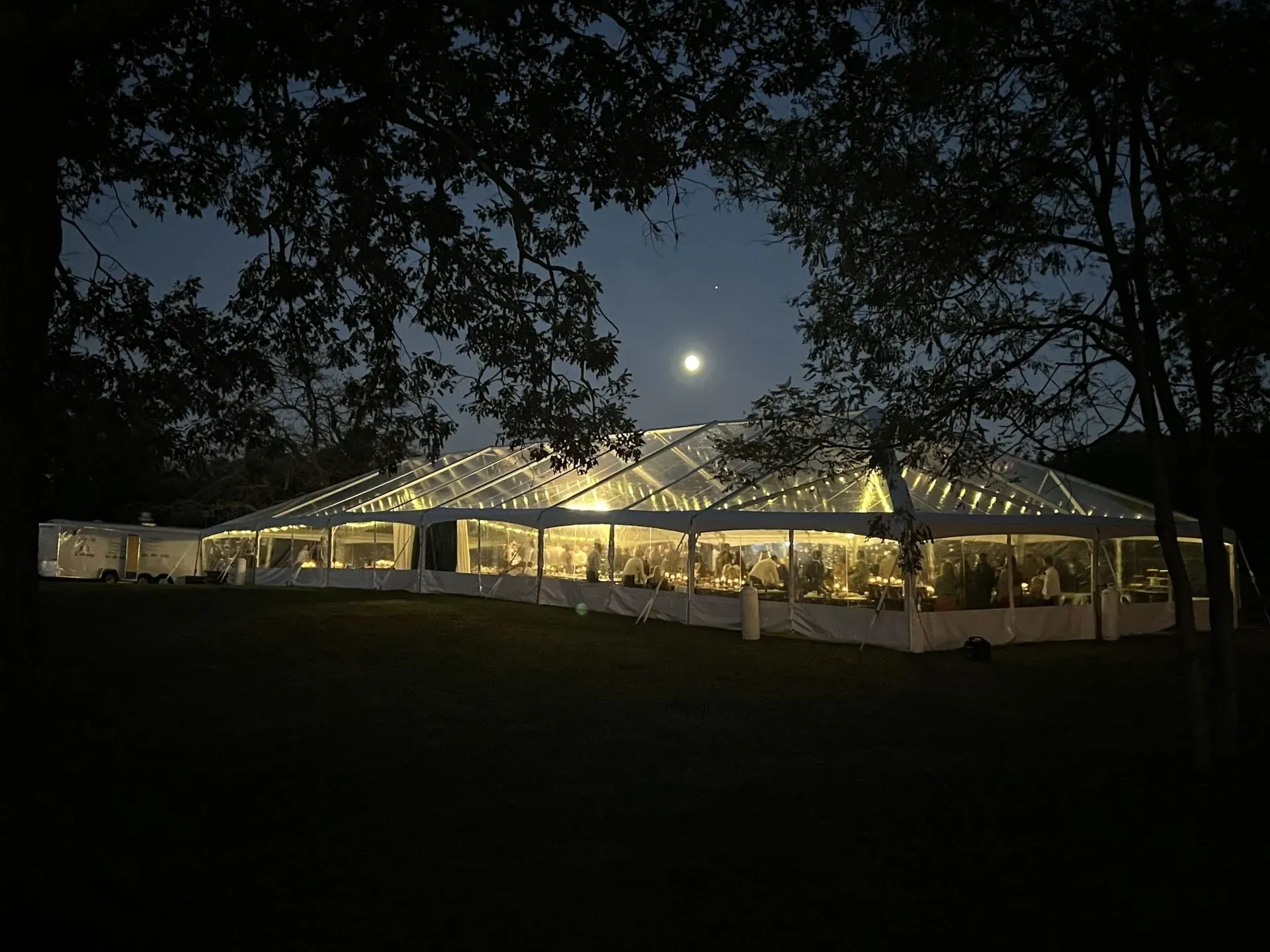 Lit clear-roof tent at night with string lights, set under a full moon, surrounded by trees.