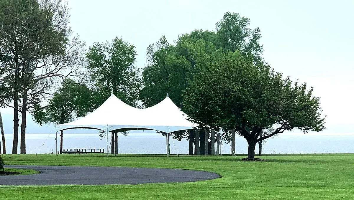 White event tent on a green lawn by a body of water with trees in the background.