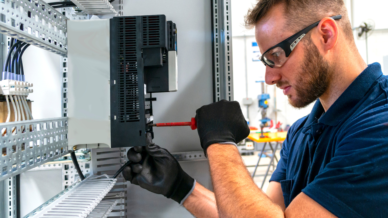 Technician in safety glasses and gloves working on electrical panel with screwdriver.