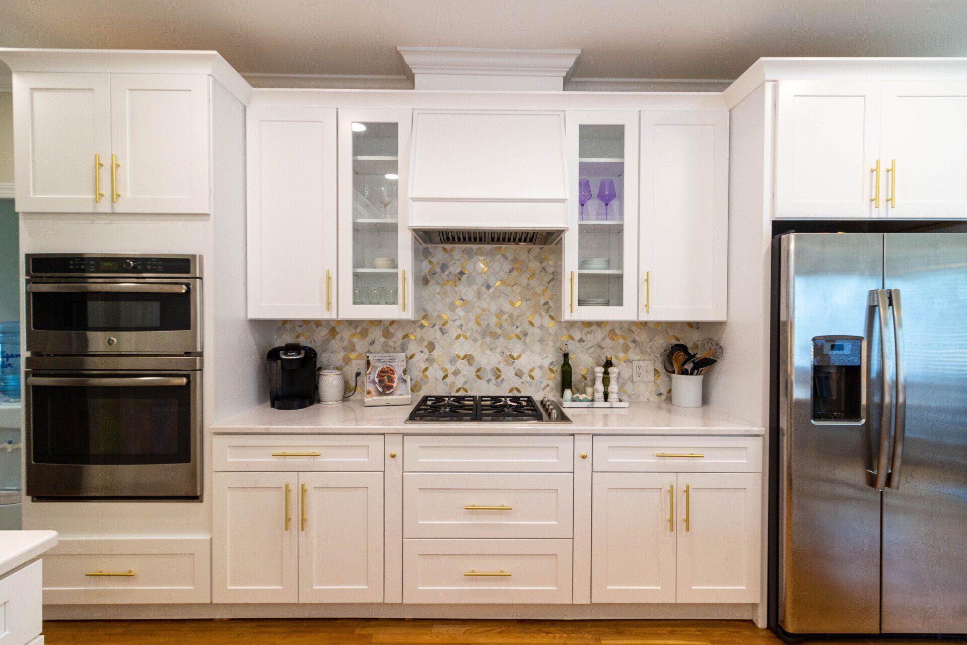Cooktop wall with custom vent hood, white cabinetry, and mosaic tile backsplash featuring gold and neutral tones.