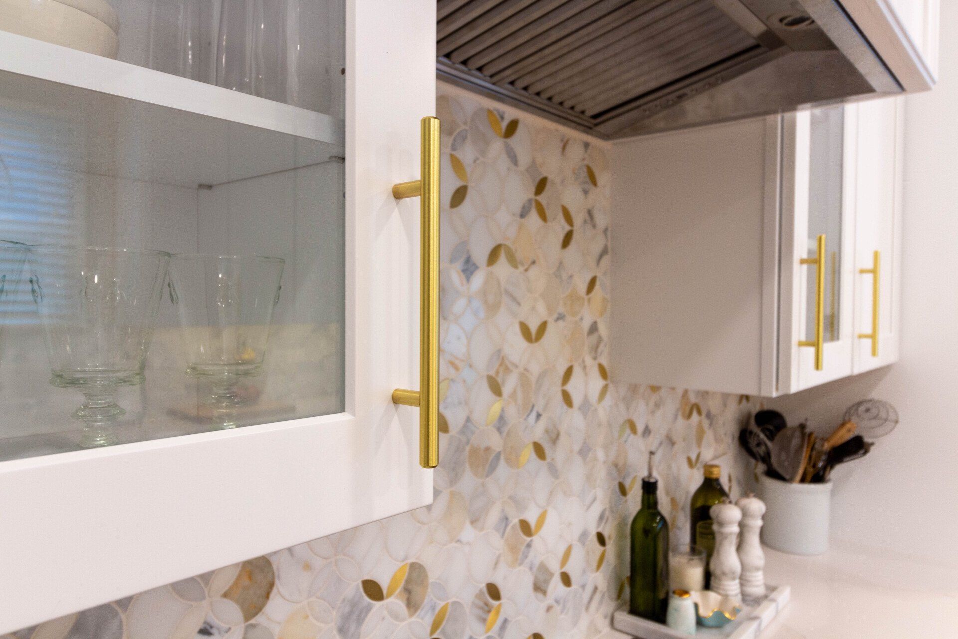Closeup of white wall cabinets with glass front and gold pull hardware on top of gold and white mosaic tile backsplash.