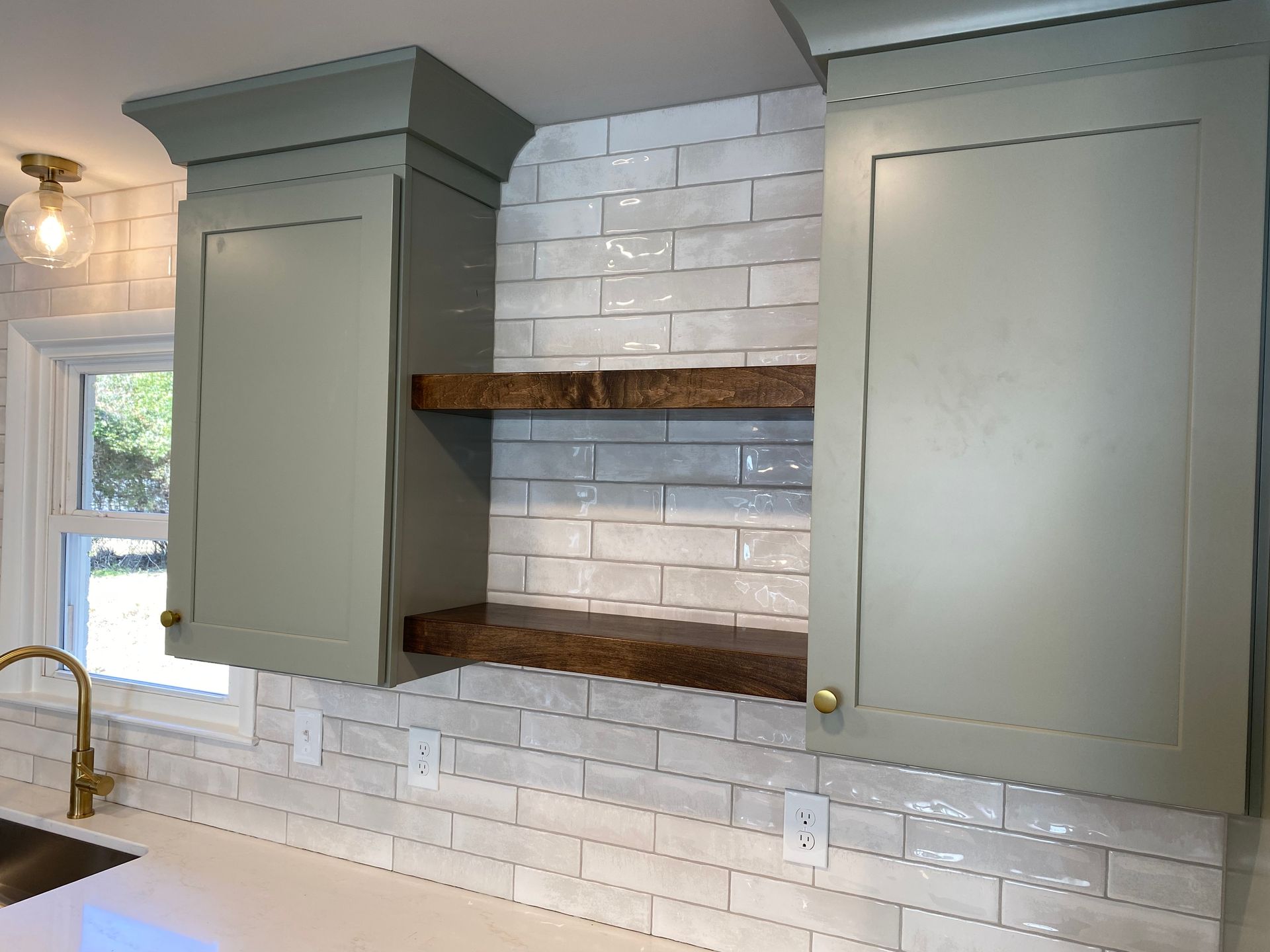 Dark wood floating shelves in between sage green shaker cabinets with white glossy subway tile backsplash.