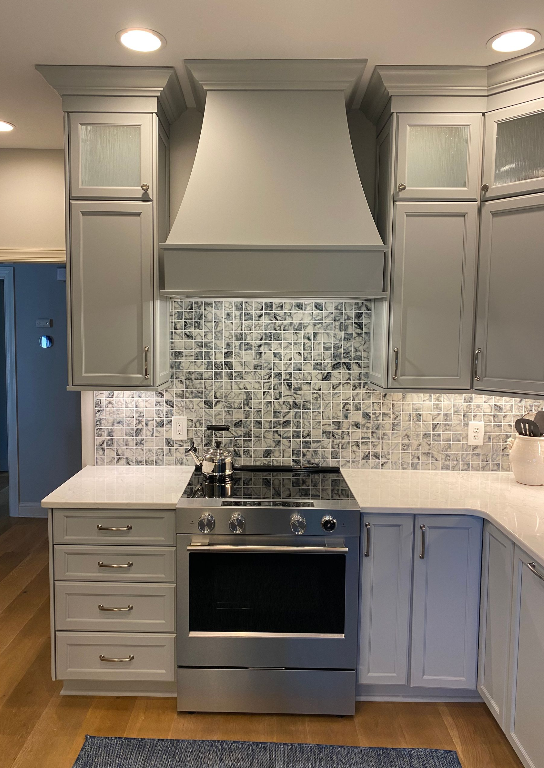 Vanguard Penta Azul mosaic tile backsplash behind gun-smoke gray custom cabinetry and matching vent hood.