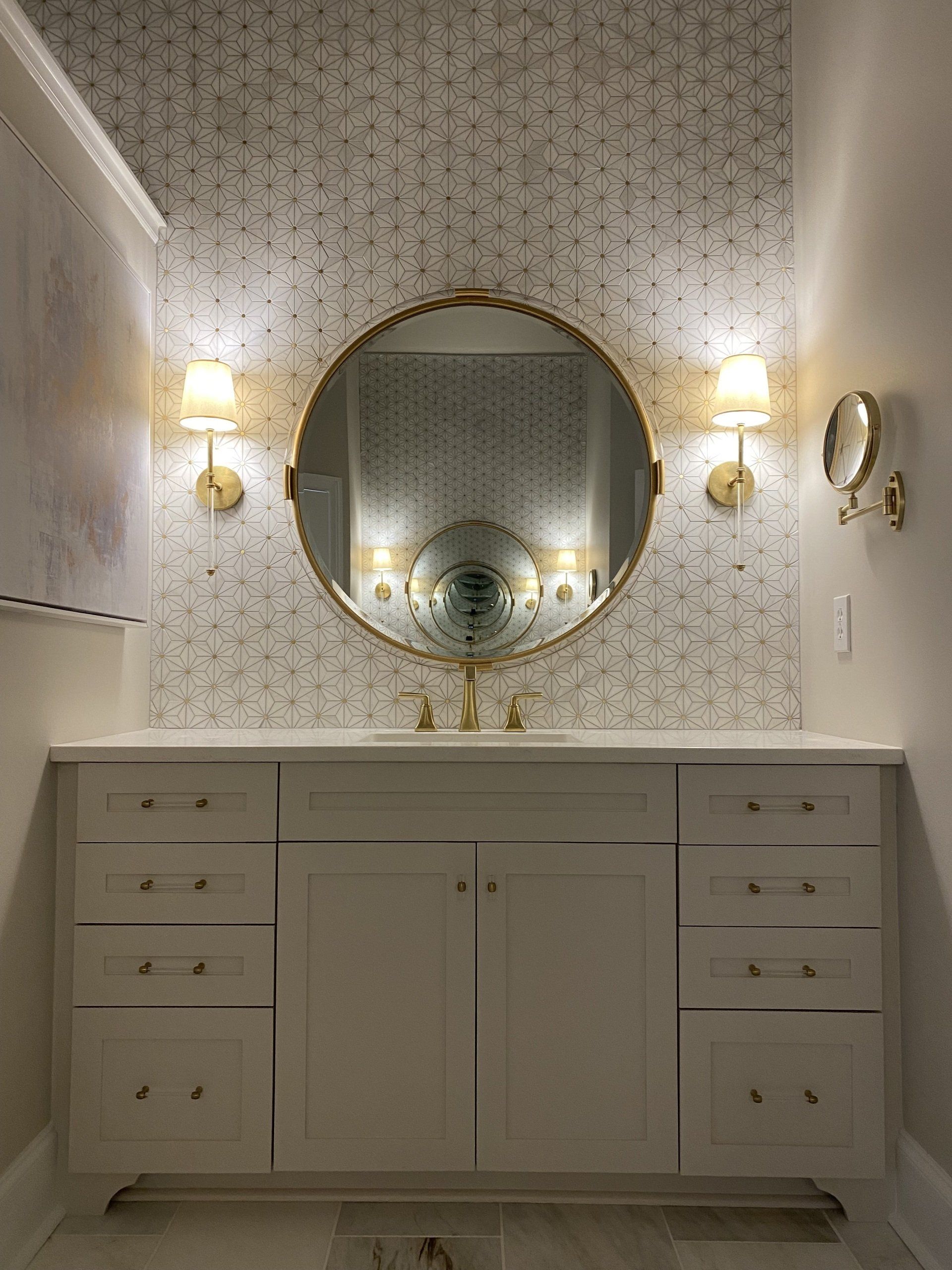 White shaker vanity with gold wall sconces, round mirror, and geometric tiled wall backsplash.