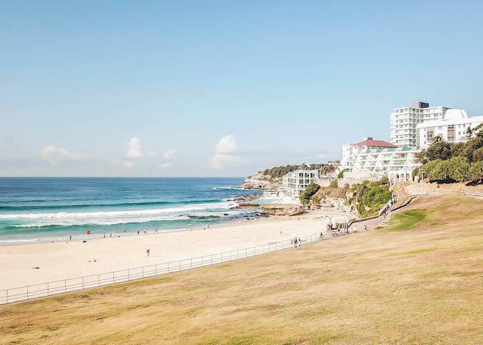 Sunny beach with turquoise waves, sandy shore, and white buildings on a grassy cliff under a clear blue sky