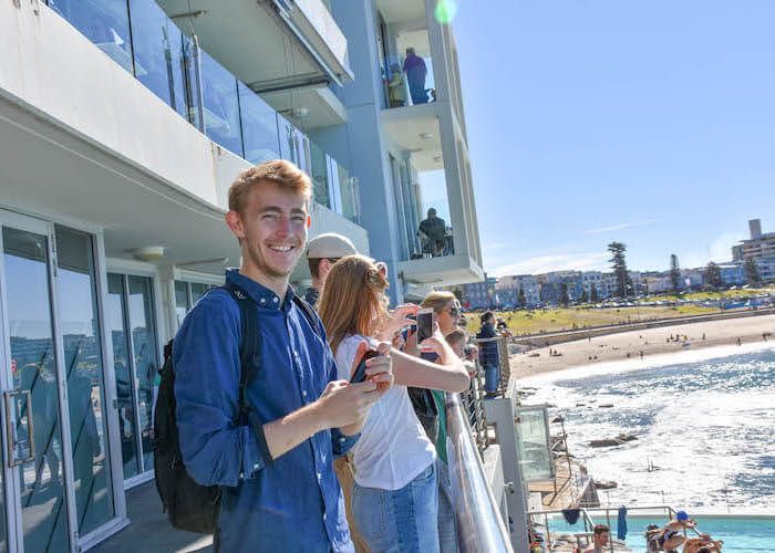 People on a sunny balcony overlooking a beach and ocean, one holding a drink and smiling.
